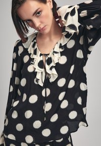 Blusa nera con grandi pois bianchi, colletto arricciato, lacci regolabili e maniche lunghe con polsini arricciati. Tessuto leggero.