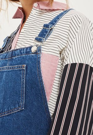 Person iført en blå denim-overalls med sølvspænder over en flerfarvet, stribet skjorte med knapper i røde, sorte og hvide mønstre.