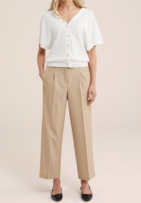 Cardigan corto bianco a maniche corte con chiusura a bottoni e scollatura a V, abbinato a pantaloni beige plissettati. Le scarpe flat nere completano l'outfit.