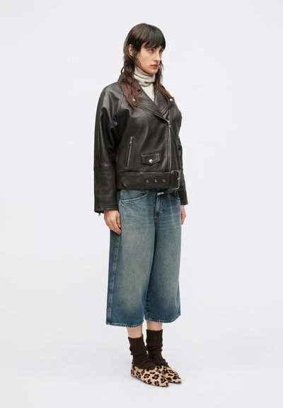 Zwarte cropped leren jas met ritsen, gecombineerd met brede blauwe jeans, bruine gebreide sokken en slip-on schoenen met luipaardprint.