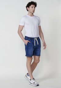 Zilas džinsu šorti ar elastīgu jostasvietu un baltu auklu, kombinēti ar baltu kreklu ar apaļu izgriezumu un baltām sporta apavām.