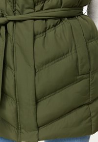 Olivgrön kviltad jacka med diagonala sömmar, sidoficka och matchande tygbälte knutet i midjan över blå jeans.
