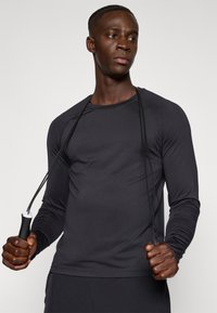 Gara piedurkņu sporta krekls tumši pelēkā krāsā, pieguloša konstrukcija, gluda tekstūra. Persona tur lecamo virvi ar melniem vadiem un baltu izcēluma rokturi.