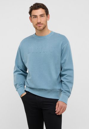 Homme portant un sweat-shirt bleu clair avec le texte en relief "MUSTANG", un pantalon noir, se tenant avec une main dans la poche devant un fond uni.