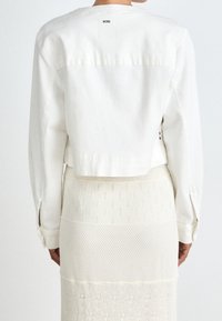 Valkoinen farkkutakki, jossa on lyhyt leikkaus ja pitkät hihat, takaosassa logotunnus. Yhdistetty valkoinen neulehame, jossa on tekstuuria.