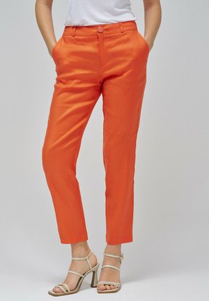 Pantalon orange en tissu lisse, avec un bouton à l'avant, des poches sur les côtés et une coupe ajustée. Porté avec des sandales à talons beiges.