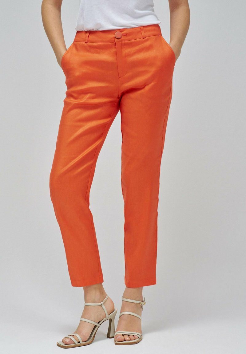 Pantalones naranja de tejido suave, con un botón en la parte delantera, bolsillos laterales y un corte entallado. Llevados con sandalias de tacón alto beige.