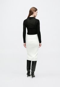 Nő, aki háttal áll, fekete hosszú ujjú felsőt, fehér, térdig érő szoknyát és fekete, magas sarkú, térdig érő csizmát visel, fehér háttéren.