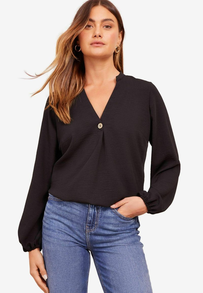 Blusa nera a maniche lunghe con scollatura a V, caratterizzata da un dettaglio centrale con bottone e una consistenza morbida. Abbinata a jeans blu in denim.