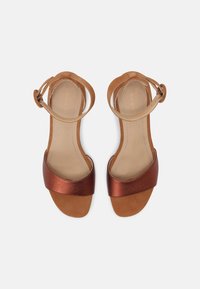 Sandales en daim marron avec des sangles en cuivre métalliques, design ouvert à l'avant et une fine bride autour de la cheville. Texture lisse et style minimaliste.