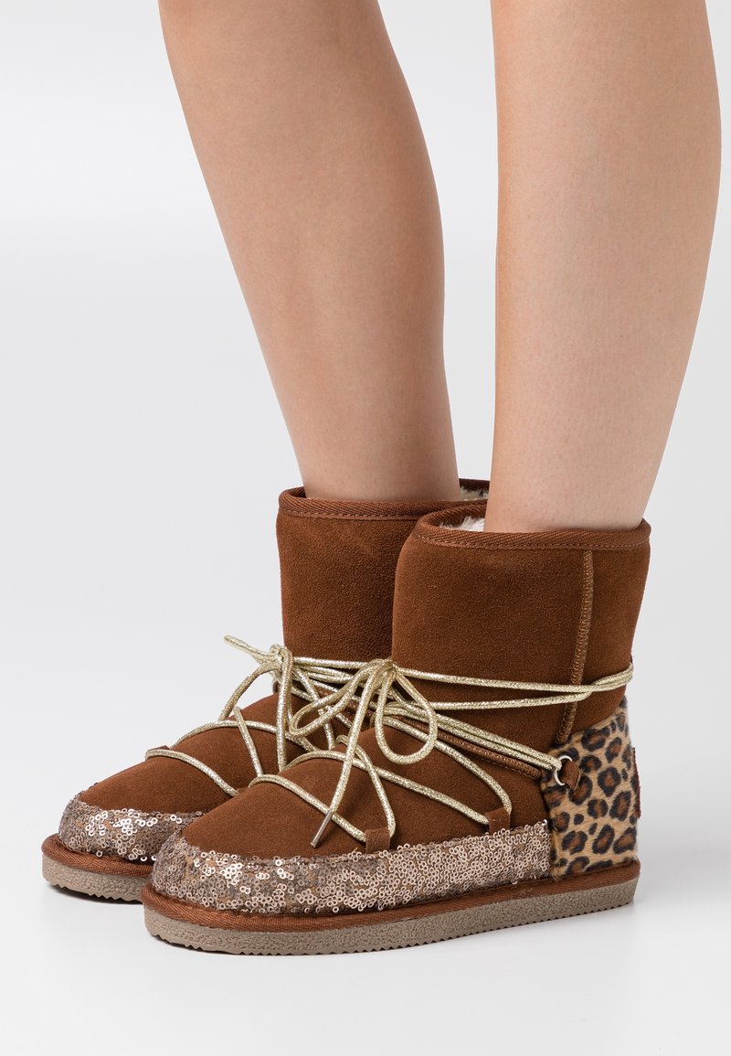 Bottines en suède marron avec lacets dorés, bouts pailletés et talons imprimés léopard, portées sur des jambes nues sur un fond blanc.