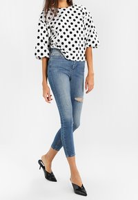 Weißes Oberteil mit schwarzen Pünktchen, Puffärmel; kombiniert mit blauen Skinny Jeans mit einem ausgefransten Knie; schwarze spitze High Heels.