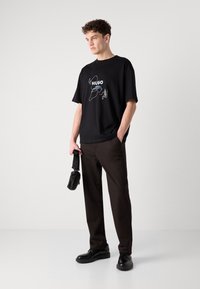 Black cotton t-shirt with white and light blue graphic design, paired with dark brown trousers and black shoes. Casual, loose fit.