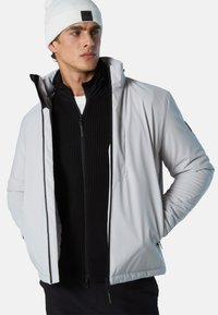 Hellgraue isolierte Jacke mit hohem Kragen, zwei Reißverschlusstaschen und schwarzen Akzenten. Trägt darunter einen schwarzen, gerippten Pullover; weiße Mütze auf dem Kopf.
