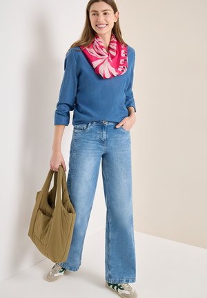 Femme en pull bleu et jean portant une écharpe rose à motifs, tenant un grand sac cabas olive, debout contre un mur beige.