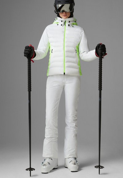 Weiße Skijacke mit neongrünen Akzenten, weiße Skihose, schwarze Handschuhe und Skistöcke. Das Model trägt einen schwarzen Helm und verspiegelte Skibrille.