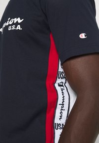 Námořnické bavlněné tričko s bílým nápisem "Champion U.S.A.", červenými bočními panely s tištěnými vzory a malým logem na rukávu.