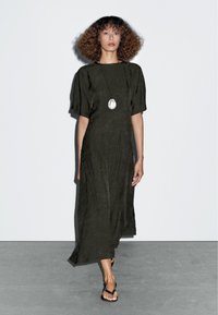 Olivegrünes knielanges Kleid aus strukturiertem Stoff, mit kurzen Ärmeln, lockerer Passform und seitlichen Schlitzen. Getragen mit schwarzen Flip-Flops.