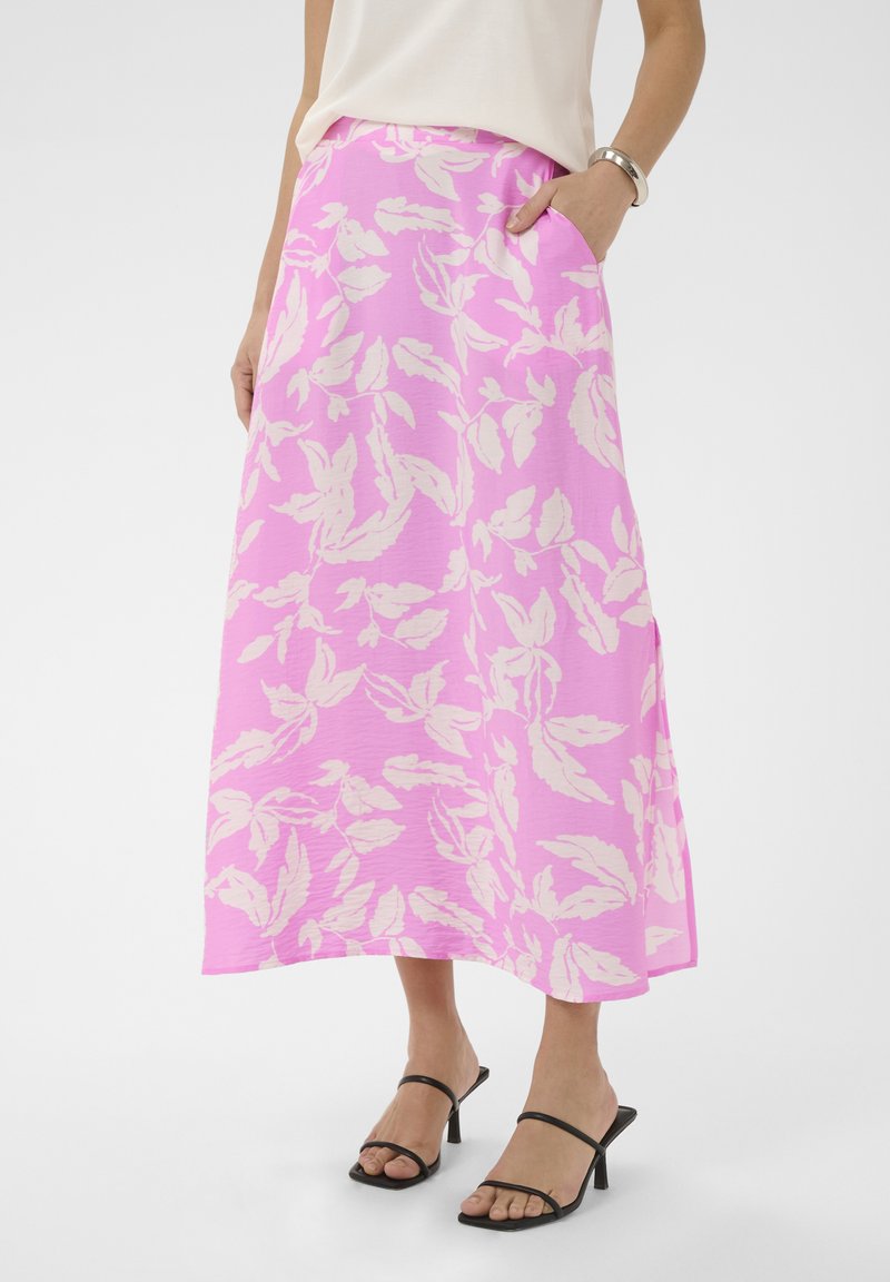 Femme portant une jupe longue rose avec des motifs de feuilles blanches, associée à des sandales à talons noires à brides et un haut blanc.