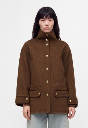 Personne portant une veste en laine marron à col haut et boutons dorés, debout devant un fond blanc uni.