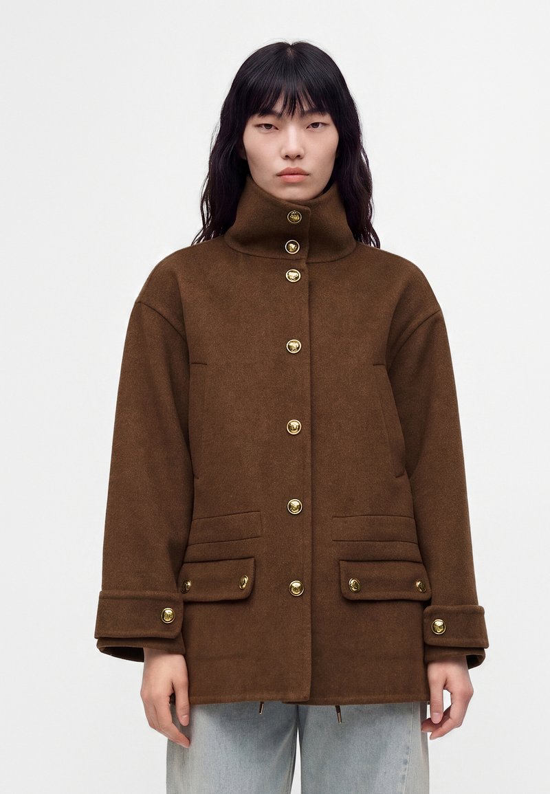 Personne portant une veste en laine marron à col haut et boutons dorés, debout devant un fond blanc uni.