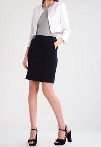 Veste blanche courte portée sur un haut gris, associée à une jupe crayon bleu marine. Sandales à talons noires complètent la tenue.