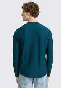 Langärmliges T-Shirt in Türkis aus strukturierter Stoff, mit Rundhalsausschnitt und lockerer Passform, kombiniert mit hellblauen Jeans.