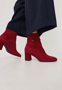 Bottines en suède rouge avec un talon carré et une fermeture éclair sur le côté, dotées d'une bande autour de la cheville et d'un accent matériel détaillé.