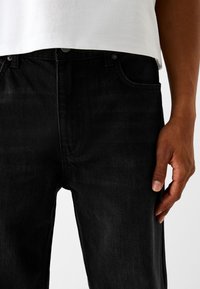 Vaqueros de mezclilla negra con un corte recto, que presentan un diseño clásico de cinco bolsillos y una textura sutil. Llevados con una camiseta blanca.
