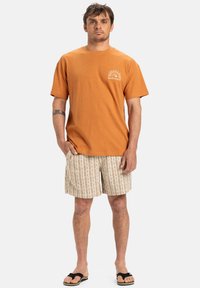 Oranžové bavlněné tričko s krátkým rukávem a logem Quiksilver. V kombinaci s béžovými pruhovanými šortkami s texturovaným vzorem a černými sandály.