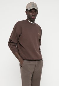 Καφέ φούτερ με στρογγυλή λαιμόκοψη και ριμπ μανσέτες, με μικρό κεντημένο λογότυπο. Φοριέται με ελαφριά γκρι κομμένα παντελόνια και μπεζ καπέλο.