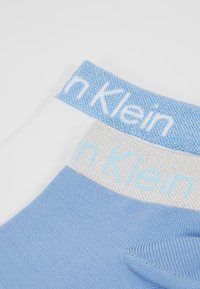 Blå og hvide sokker med et struktureret lyseblåt bånd, der har "Calvin Klein" logoet. Glat stof med en kontrasterende sølvsektion.