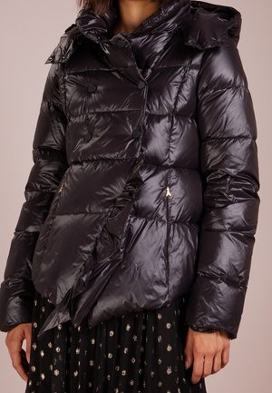 Błyszcząca czarna pikowana kurtka puchowa z dużym kołnierzem, podwójnymi czarnymi guzikami i zapinanymi na zamek bocznymi kieszeniami, noszona na czarnej spódnicy.