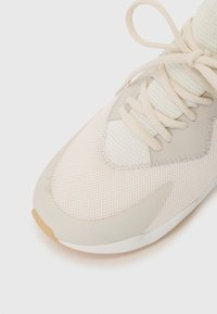 Lys beige treningssko med mesh-overdel, gummisåle og polstret tunge. Snøring og glatte detaljer nær tåen og sidene.