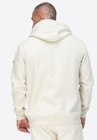 Sudadera color crema con un acabado texturizado, que presenta una gran capucha, puños y dobladillo acanalados. Incluye un pequeño logo en la manga izquierda.