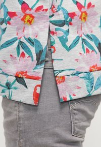 Top de manga corta floral en gris claro con flores vibrantes en rosa y verde azulado, con una abertura lateral. Combinado con pantalones grises.
