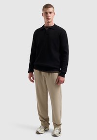 Jonge man die staat in een zwarte longsleeve poloshirt, beige broek en witte sneakers met patroon tegen een effen lichtgrijze achtergrond.