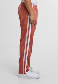 Pfirsichfarbene Jogginghose mit weißen Seitenstreifen, elastischem Bund und Reißverschlüssen an den Bündchen. Hergestellt aus glattem Stoff mit einer entspannten Passform.