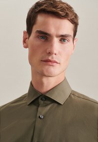Olijfgroene shirt met een gestructureerde kraag, met een gladde textuur en twee donkere knopen aan de voorkant. Close-up van de halslijn van het kledingstuk.