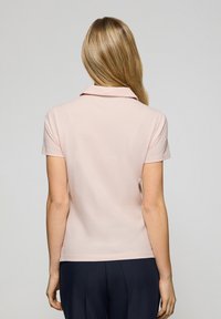 Polo shirt rosa chiaro con colletto, maniche corte e texture morbida, visto da dietro su uno sfondo neutro.