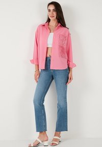 Pink button-up shirt, ribbed texture, features a lace pocket detail. Worn with light blue flared jeans and white strappy sandals.