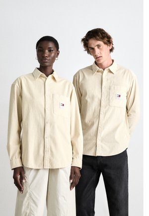 Dos personas visten camisas de botonadura en corduroy de color beige claro con bolsillos en el pecho; una lleva pantalones claros y la otra jeans oscuros.