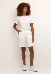 T-shirt bianco in cotone abbinato a pantaloni corti bianchi fino al ginocchio, con tasche laterali, tessuto morbido e cintura elastica. Scarpe da ginnastica bianche completano il look.