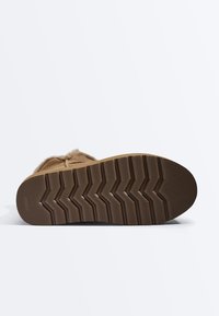 Semelle de botte marron avec un motif de bande de roulement en zigzag profond et des sections texturées, montrant un design antidérapant et une partie de la tige douce doublée de fourrure.