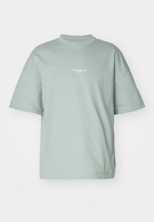 T-shirt oversized vert clair en coton doux, avec des manches courtes et un col rond, portant l'inscription "ORIGINALS est. 1990" en blanc.