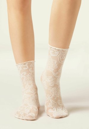 Voeten met doorschijnende witte kanten sokken met bloemenpatronen, staand op een neutrale achtergrond.