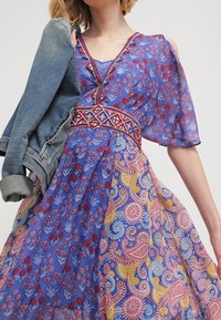 Modré květinové šaty s paisley vzory, průsvitná látka a výstřih do V. Detaily zahrnují červenou výšivku a džínovou bundu.