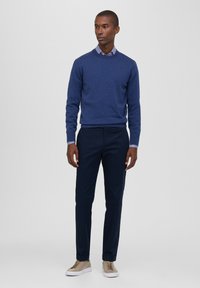 Maglione blu navy sopra una camicia a righe, abbinato a pantaloni blu navy e sneakers beige. L'outfit presenta un design semplice e casual.