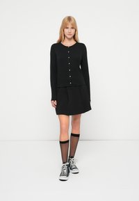 Cárdigan negro de manga larga con cierre de botones, combinado con una falda plisada negra, calcetas negras hasta la rodilla y zapatillas negras.