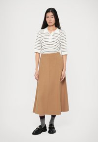 Weißes Kurzarmoberteil mit bunten Streifen, kombiniert mit einem langen, camel-farbenen A-Linien-Rock. Das Model trägt schwarze Schuhe und graue Socken.